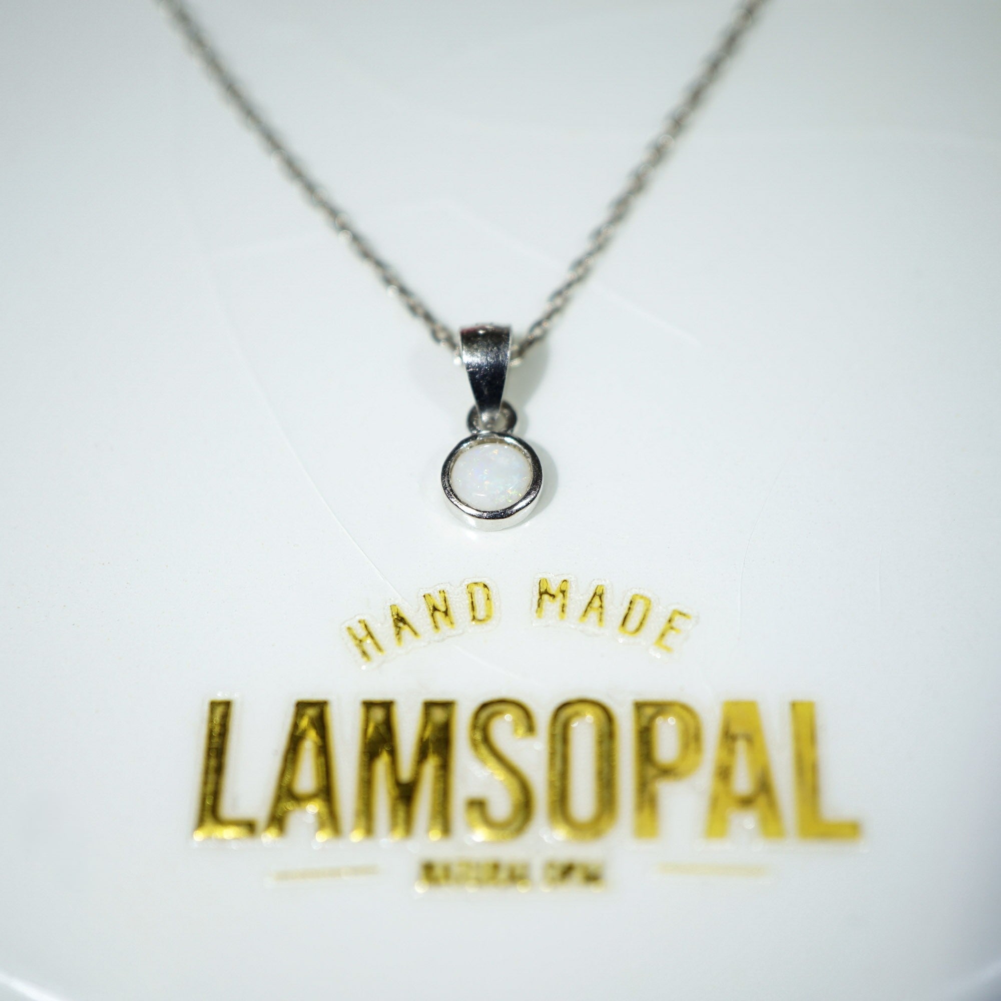 A silver chain necklace with a small circular pendant on a white display card. The card features gold foil text reading 'HAND MADE LAMSOPAL'.