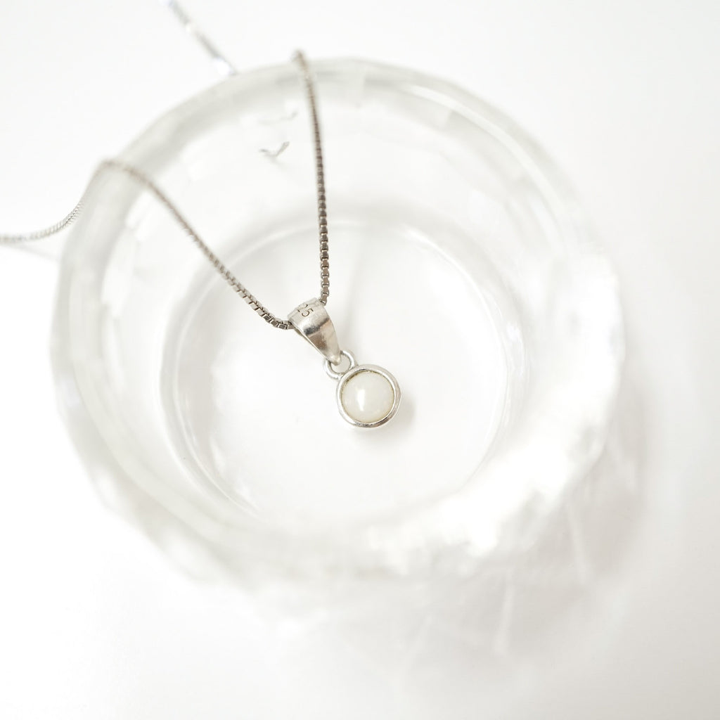 A silver chain necklace with a small oval pendant featuring a milky white stone, displayed inside a clear glass dish on a white background.