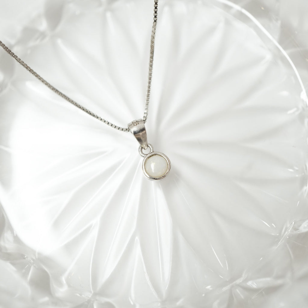 A silver chain necklace with a small circular pendant featuring a milky white stone, displayed on a white textured fabric background.
