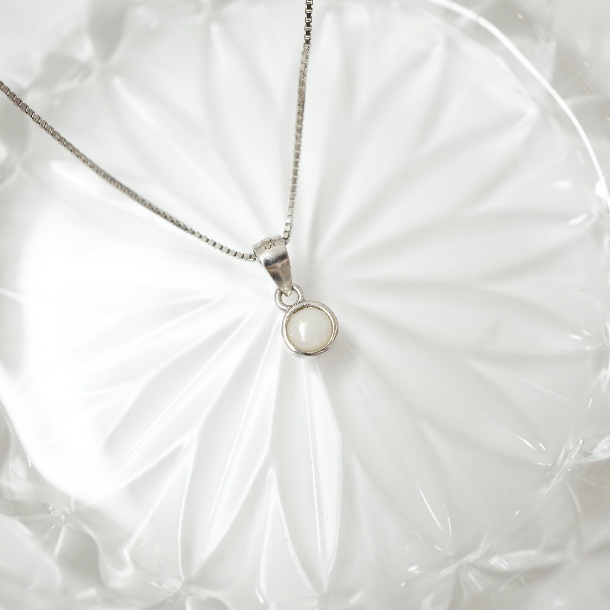 A silver chain necklace with a small circular pendant featuring a milky white stone, displayed on a white textured fabric background.