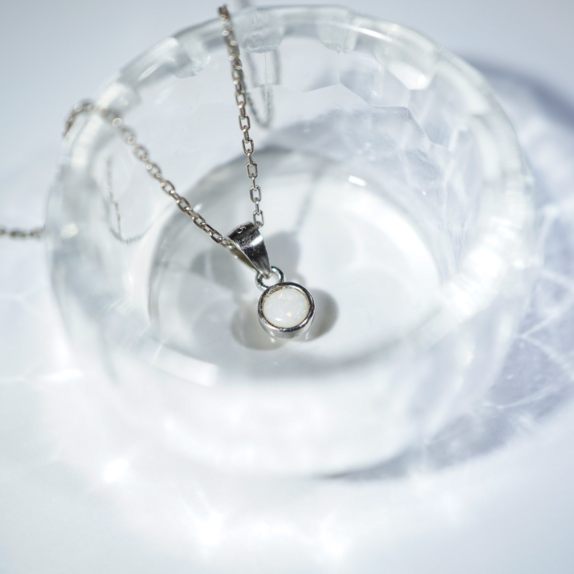 A silver chain necklace with a minimalist circular ring pendant, displayed inside a clear faceted glass bowl on a white surface.