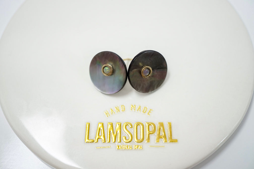 A pair of round stud earrings with iridescent mother-of-pearl discs and small gold-toned bezel-set stones, displayed on a white circular branded card that reads LAMSOPAL.