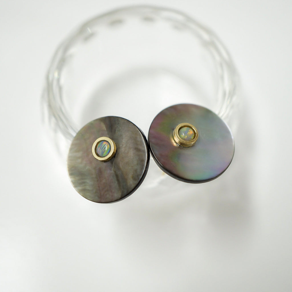 A pair of round stud earrings with iridescent mother-of-pearl disc faces and gold bezel-set iridescent centers, displayed on a clear glass stand against a white background.