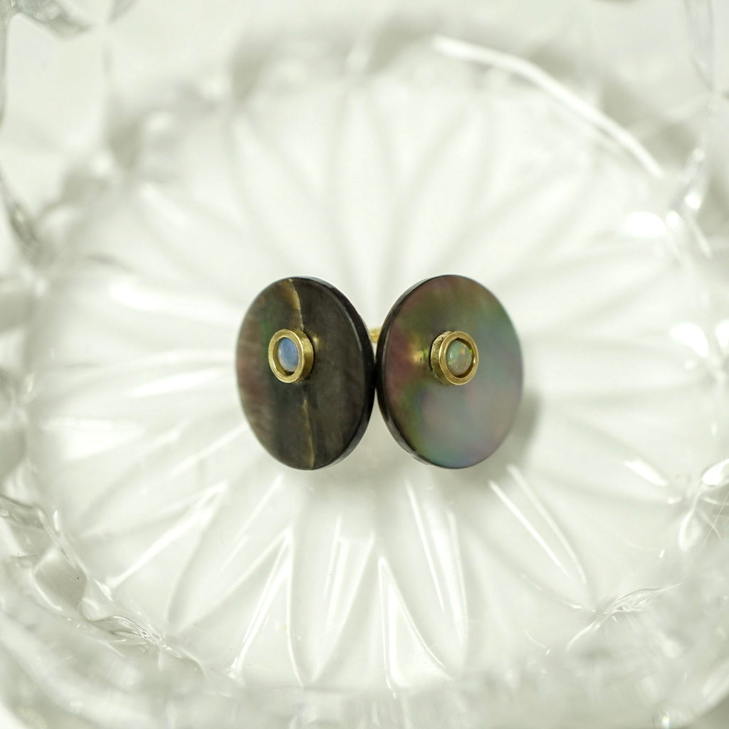 A pair of round stud earrings with iridescent mother-of-pearl discs and small gold bezel-set blue stones, displayed on a textured clear glass dish.