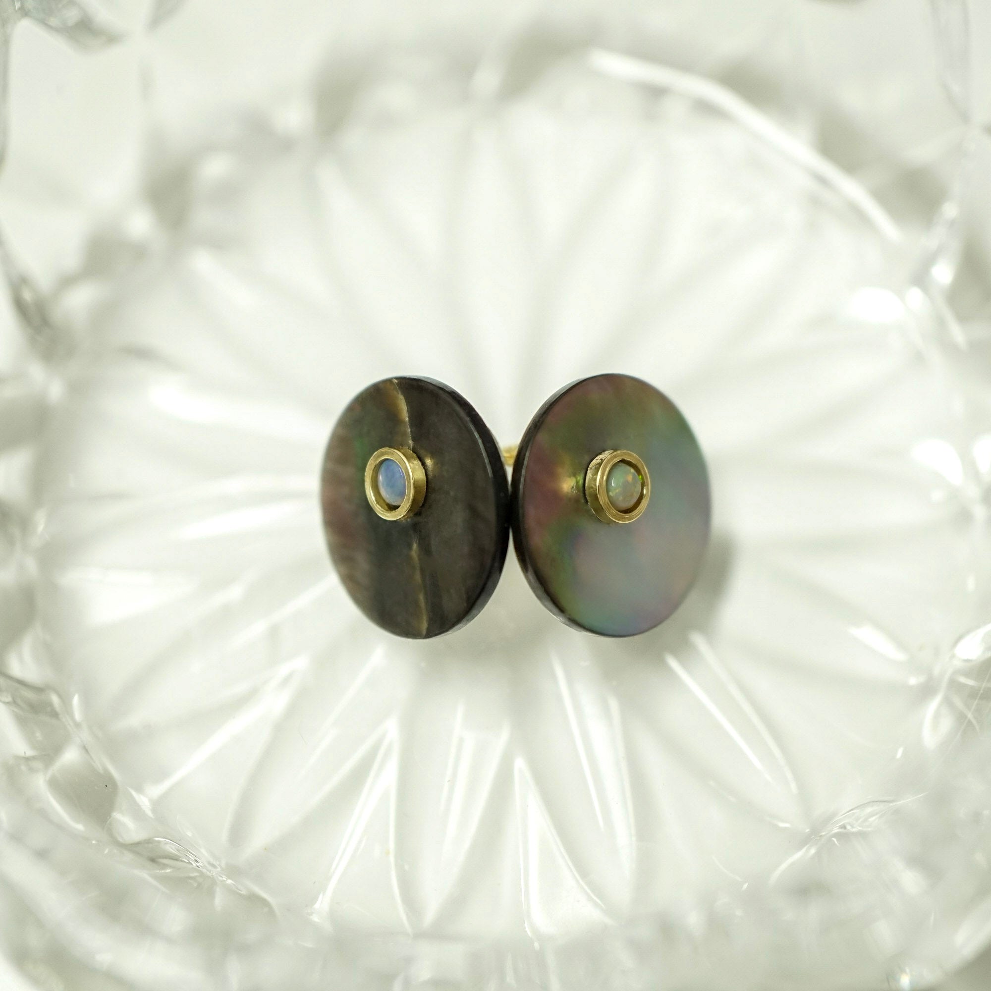 A pair of round stud earrings with iridescent mother-of-pearl discs and small gold bezel-set blue stones, displayed on a textured clear glass dish.
