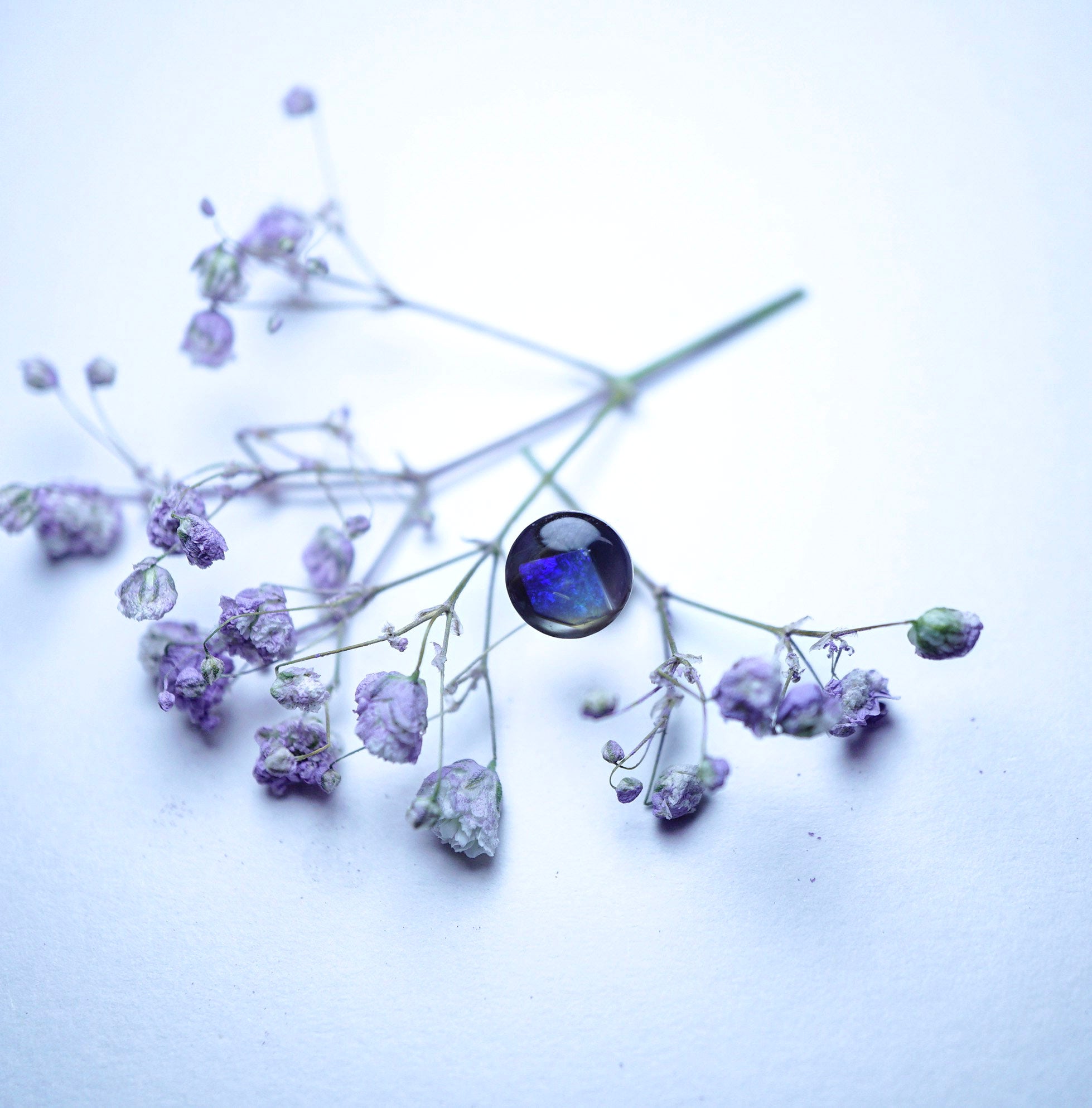 A delicate floral arrangement with small pale purple blossoms and thin stems on a soft white background, featuring a prominent round dark blue gemstone accent.