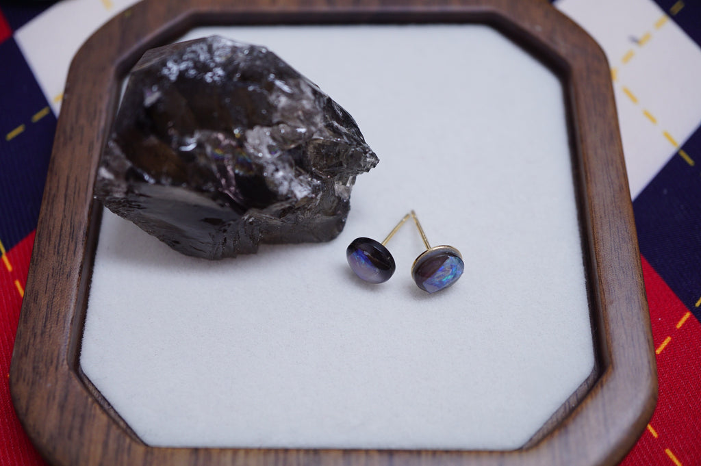 A pair of oval stud earrings with dark brown oval stones and gold posts, displayed on a white insert inside a wooden octagonal tray next to a dark mineral specimen.