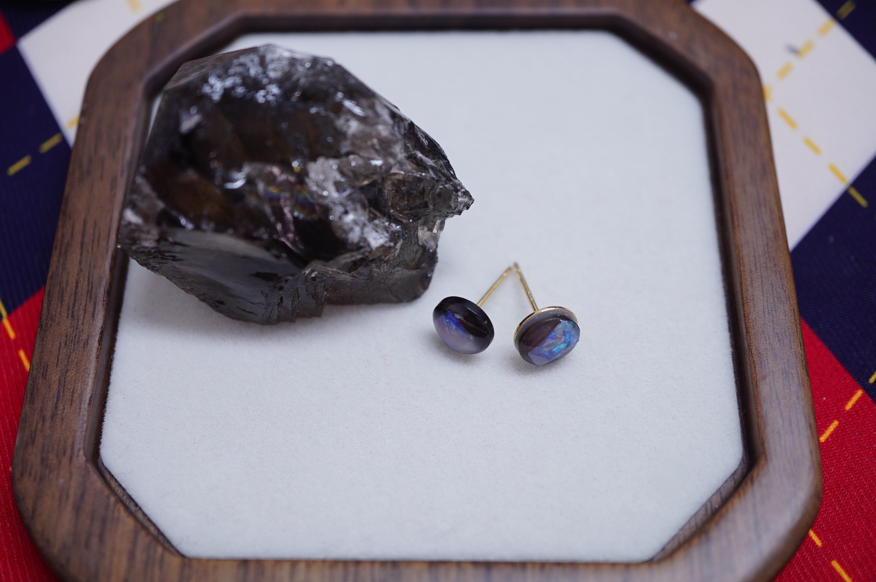A pair of oval stud earrings with dark brown oval stones and gold posts, displayed on a white insert inside a wooden octagonal tray next to a dark mineral specimen.