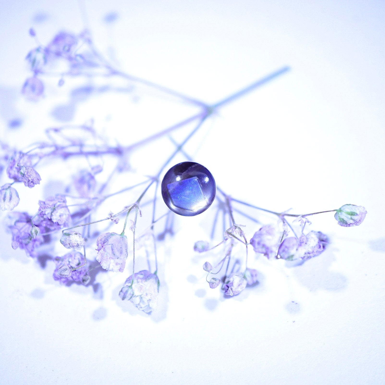 A close-up of delicate pale purple dried flower sprigs with a single glossy oval blue-purple gemstone or bead at the center, set against a bright white background with soft light leaks.