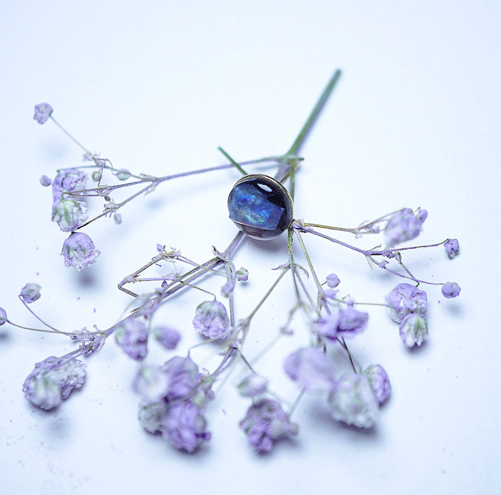 A small floral arrangement with pale lavender dried blossoms and stems, centered around a dark oval object with a blue reflective center, photographed on a plain white background.