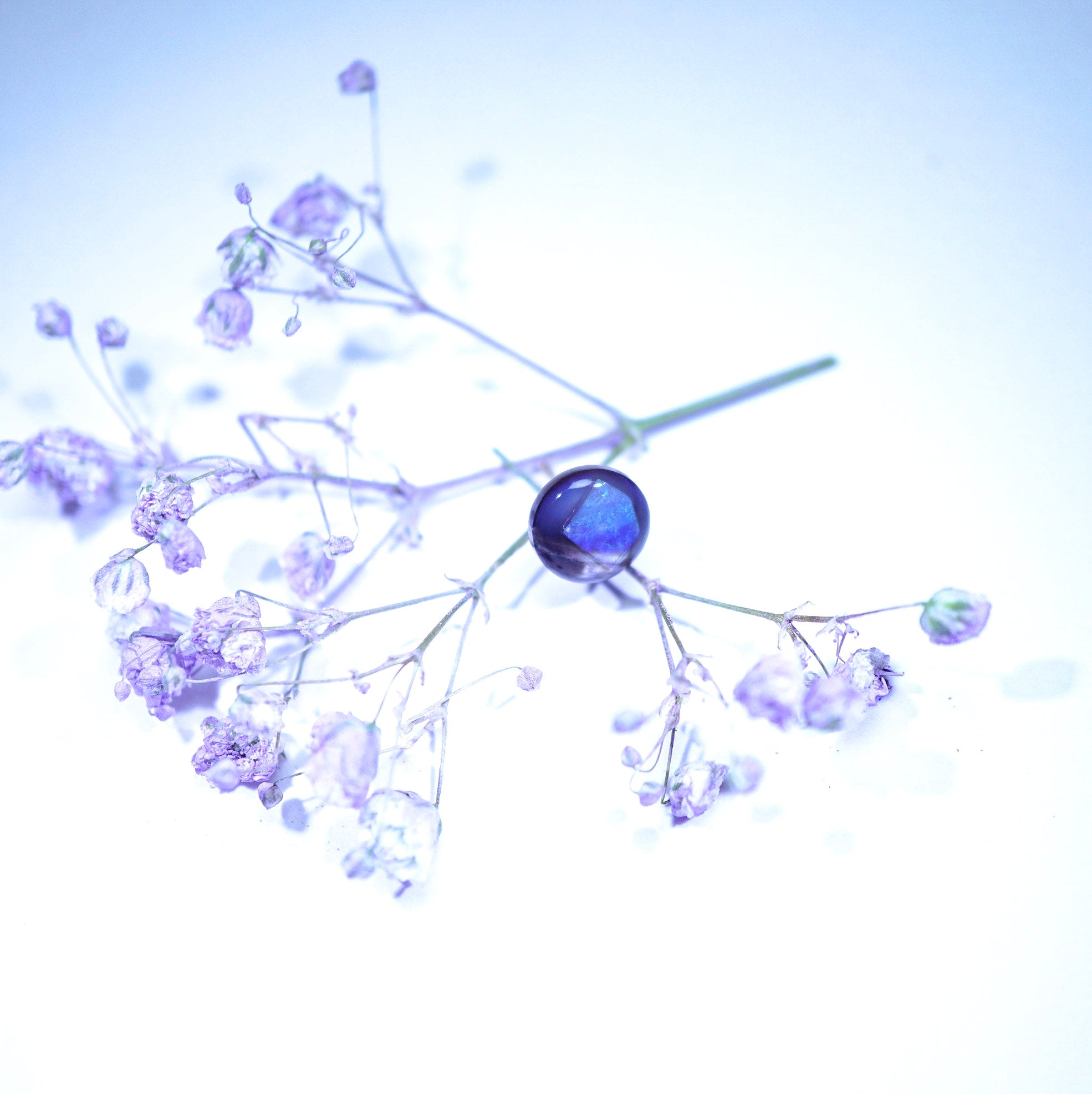 A delicate dried floral arrangement with pale lavender sprigs and a central glossy dark blue oval gemstone, photographed against a bright white background with a cool blue tint.