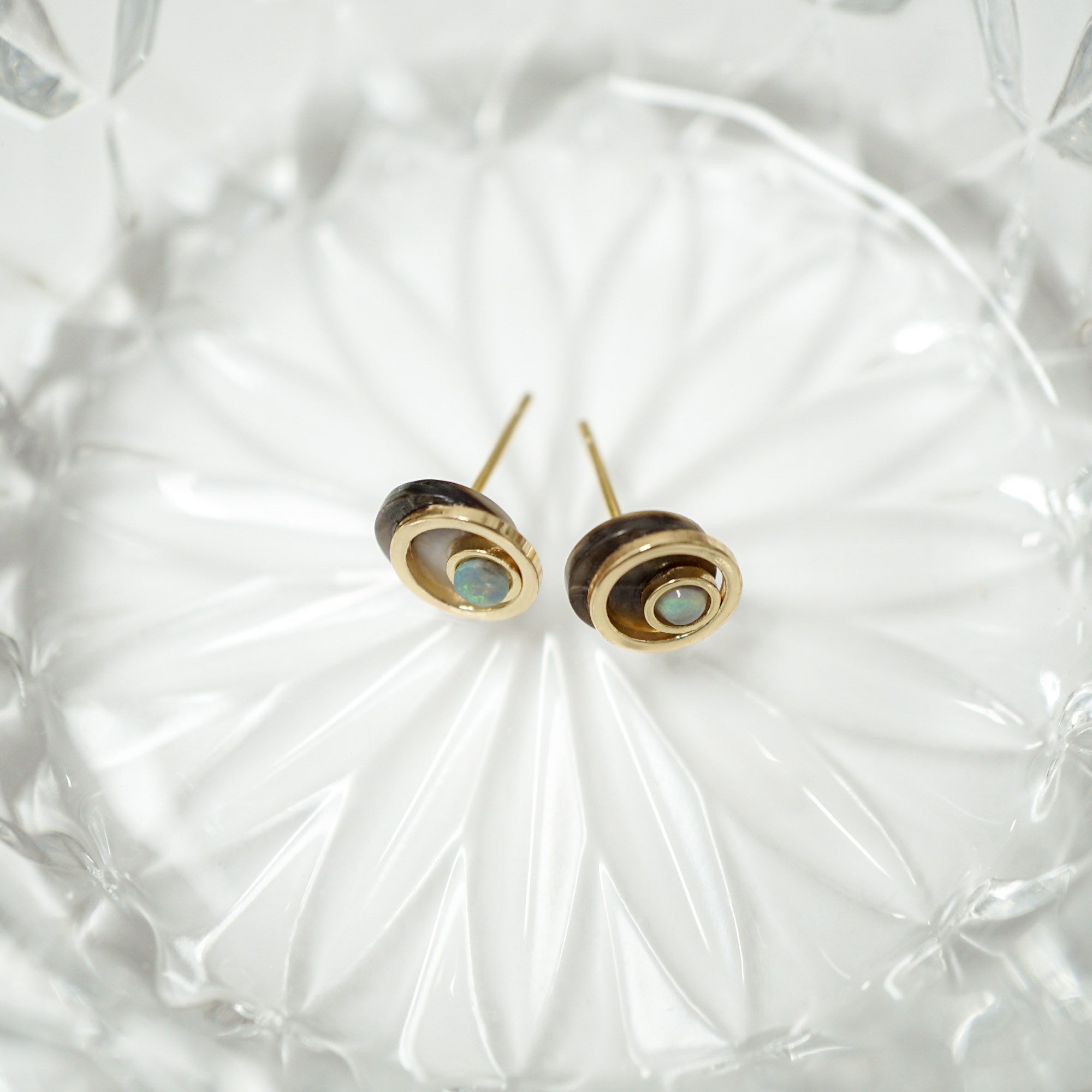 A pair of gold and dark brown stacked disc stud earrings with oval light blue-green inlays, resting on a textured clear glass dish.