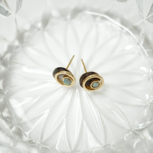 A pair of gold and dark brown stacked disc stud earrings with oval light blue-green inlays, resting on a textured clear glass dish.