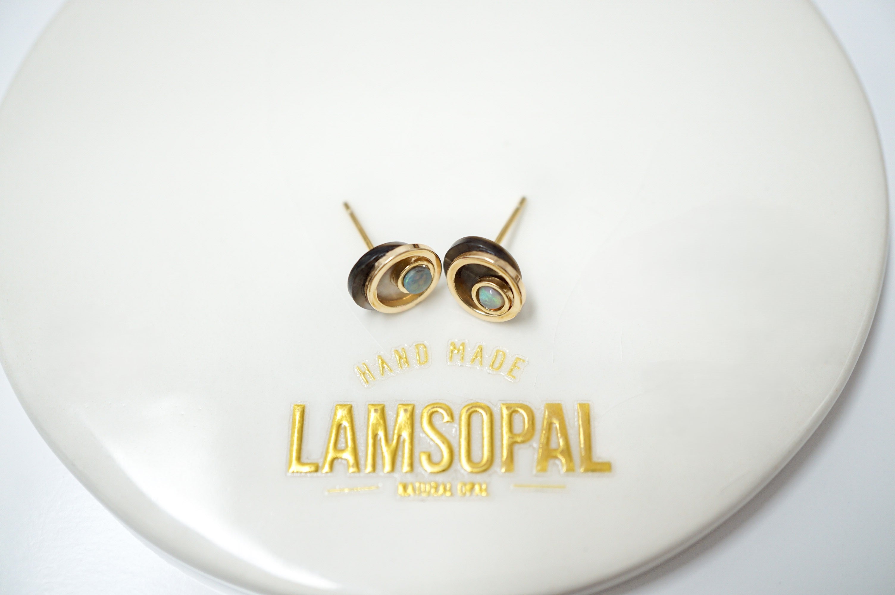 A pair of gold-toned spiral stud earrings with light blue-green stone centers, displayed on a white circular plate printed with the brand name 'LAMSOPAL'.