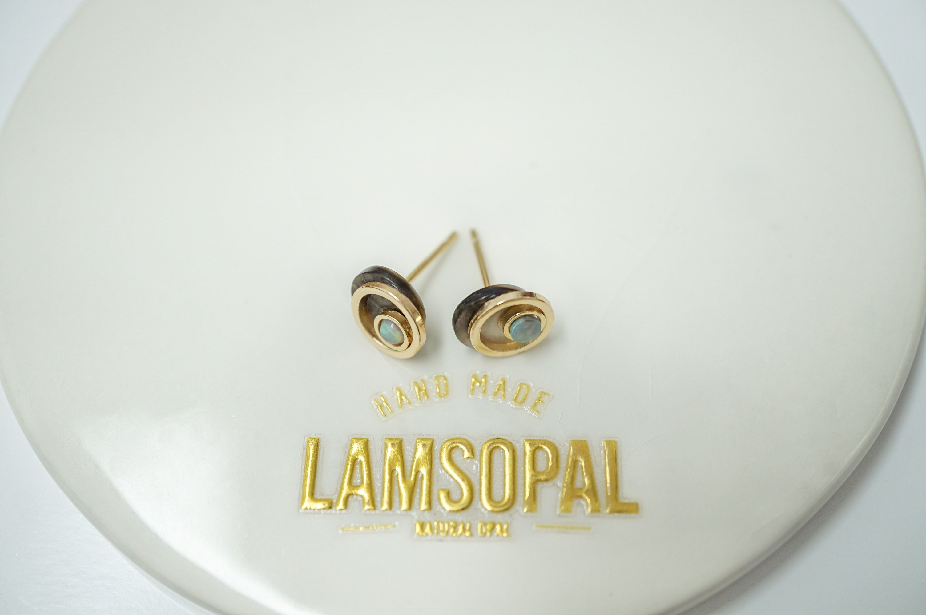 A pair of small oval stud earrings displayed on a white circular stand. The earrings have a gold-tone outer ring, a dark brown center, and a light blue-green stone in the middle.