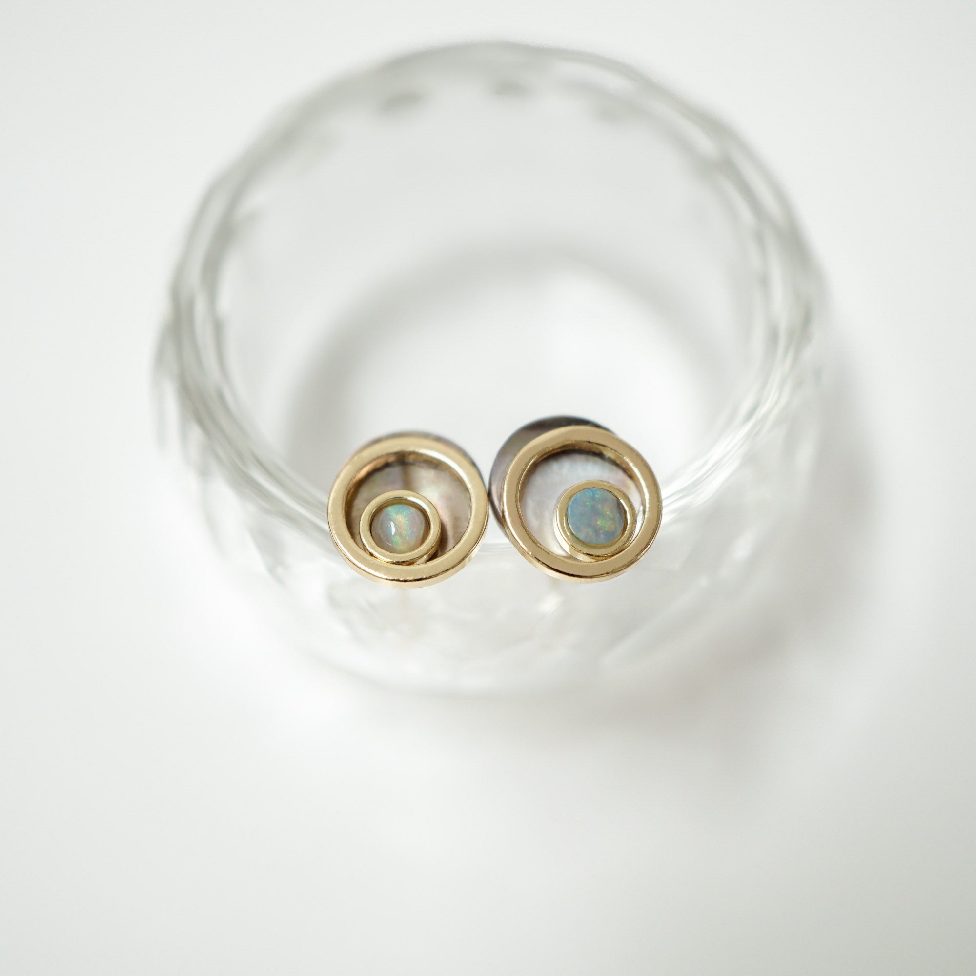 A pair of gold-toned spiral stud earrings with small iridescent blue-green opal centers, displayed on a clear glass coaster against a white background.
