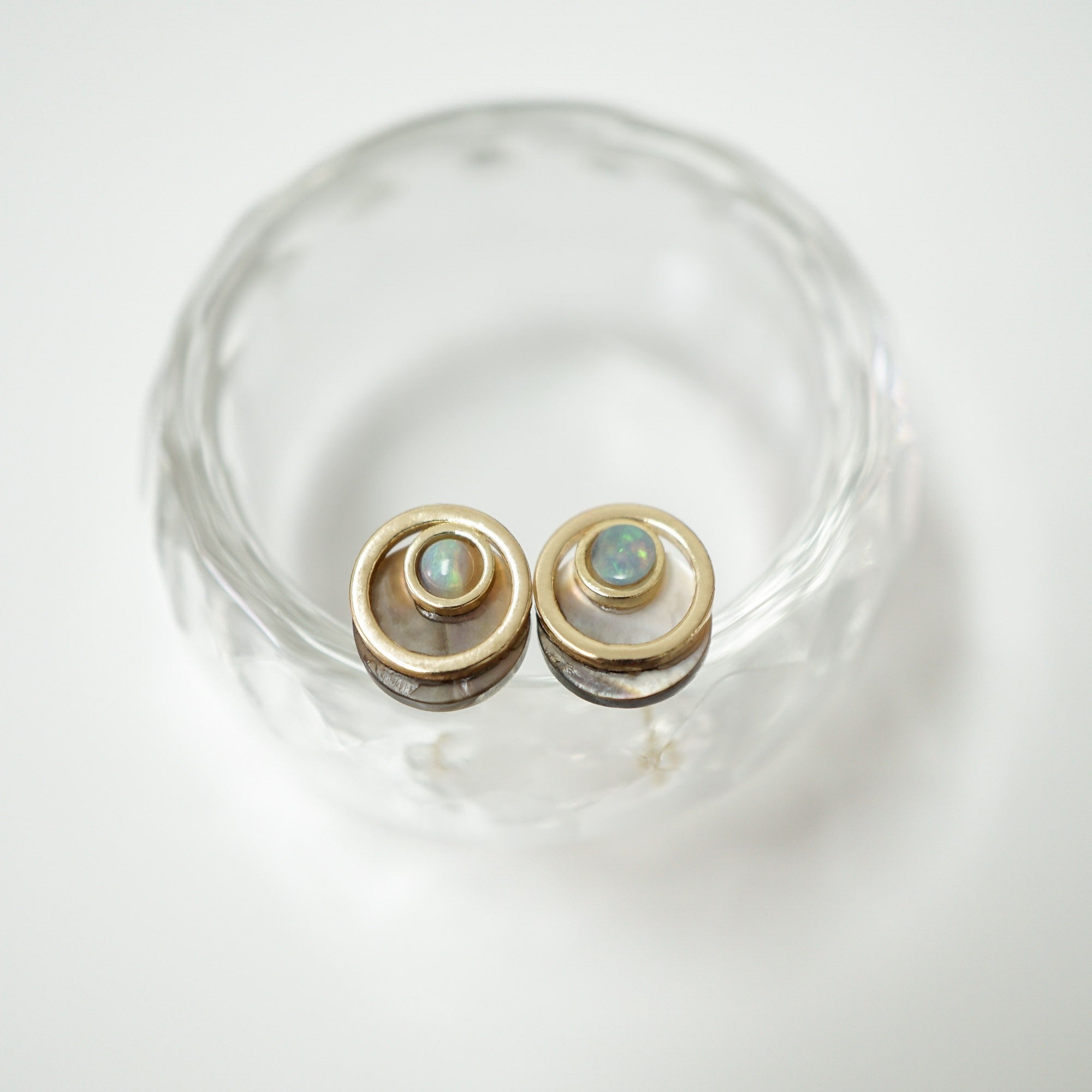 A pair of gold-toned spiral stud earrings with small iridescent blue-green opal-like centers, displayed on a clear glass dish against a white background.