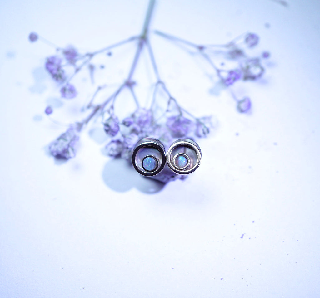 A pair of silver-toned hoop earrings with oval iridescent blue-white centers, resting on delicate dried lavender sprigs against a pale lavender background.