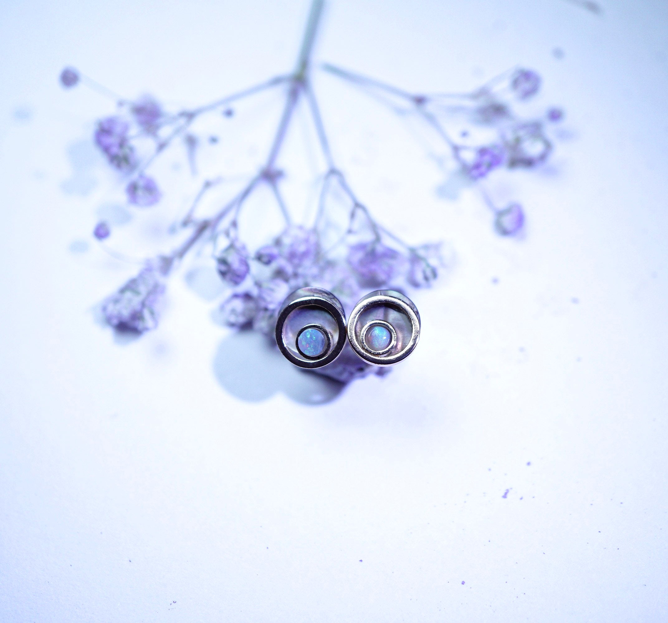 A pair of silver-toned hoop earrings with oval iridescent blue-white centers, resting on delicate dried lavender sprigs against a pale lavender background.