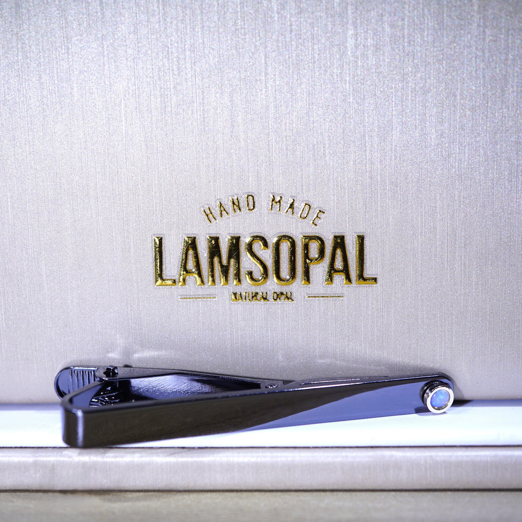 A black metal nail clipper with a blue accent ring, resting on a white surface in front of a textured light gray background with gold 'LAMSOPAL' branding.