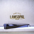 A black metal nail clipper with a blue accent ring, resting on a white surface in front of a textured light gray background with gold 'LAMSOPAL' branding.