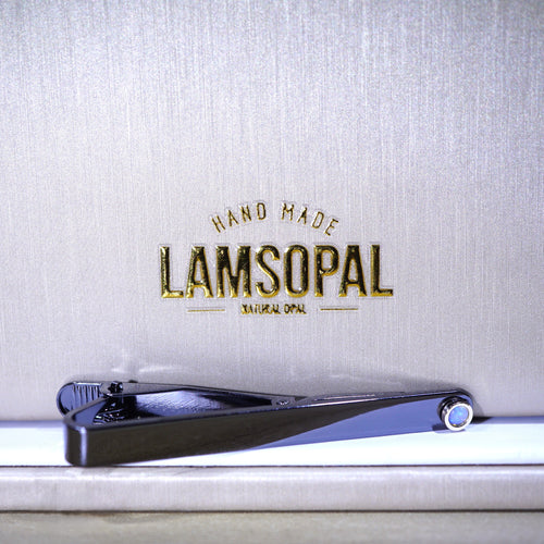 A black metal nail clipper with a blue accent ring, resting on a white surface in front of a textured light gray background with gold 'LAMSOPAL' branding.