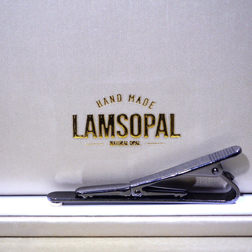 A close-up of a black metal hair clip resting on a white surface in front of a textured light gray background with the brand name 'LAMSOPAL' printed in gold and black.