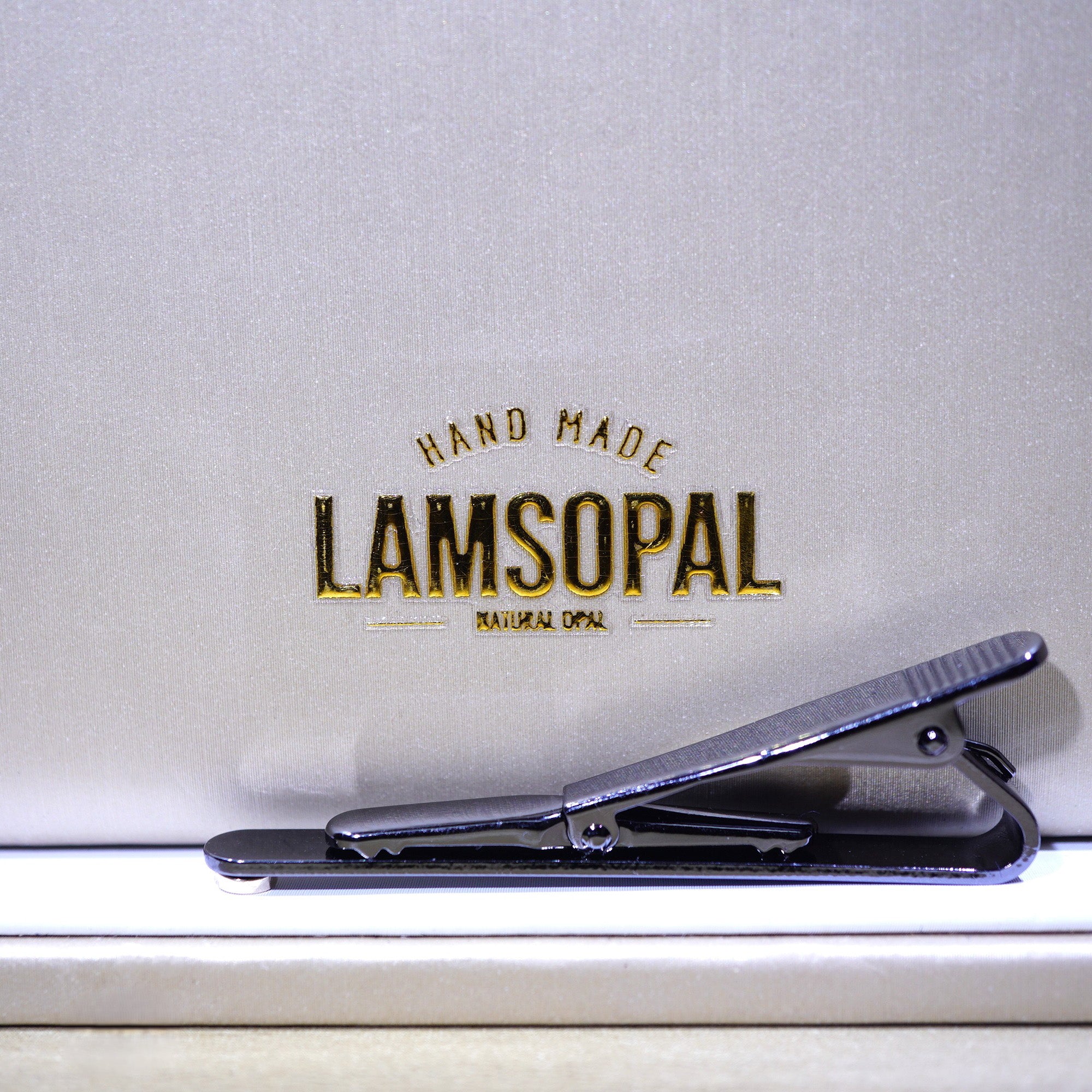 A close-up of a black metal hair clip resting on a white surface in front of a textured light gray background with the brand name 'LAMSOPAL' printed in gold and black.