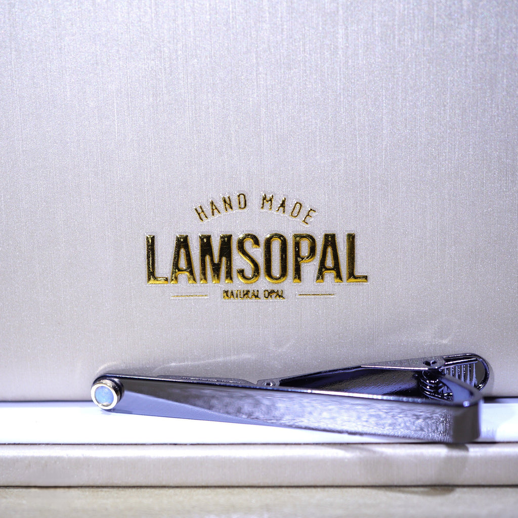 A silver metal tie clip with a textured finish, resting on a white surface in front of a textured light gray background with gold foil branding that reads 'LAMSOPAL HAND MADE'.