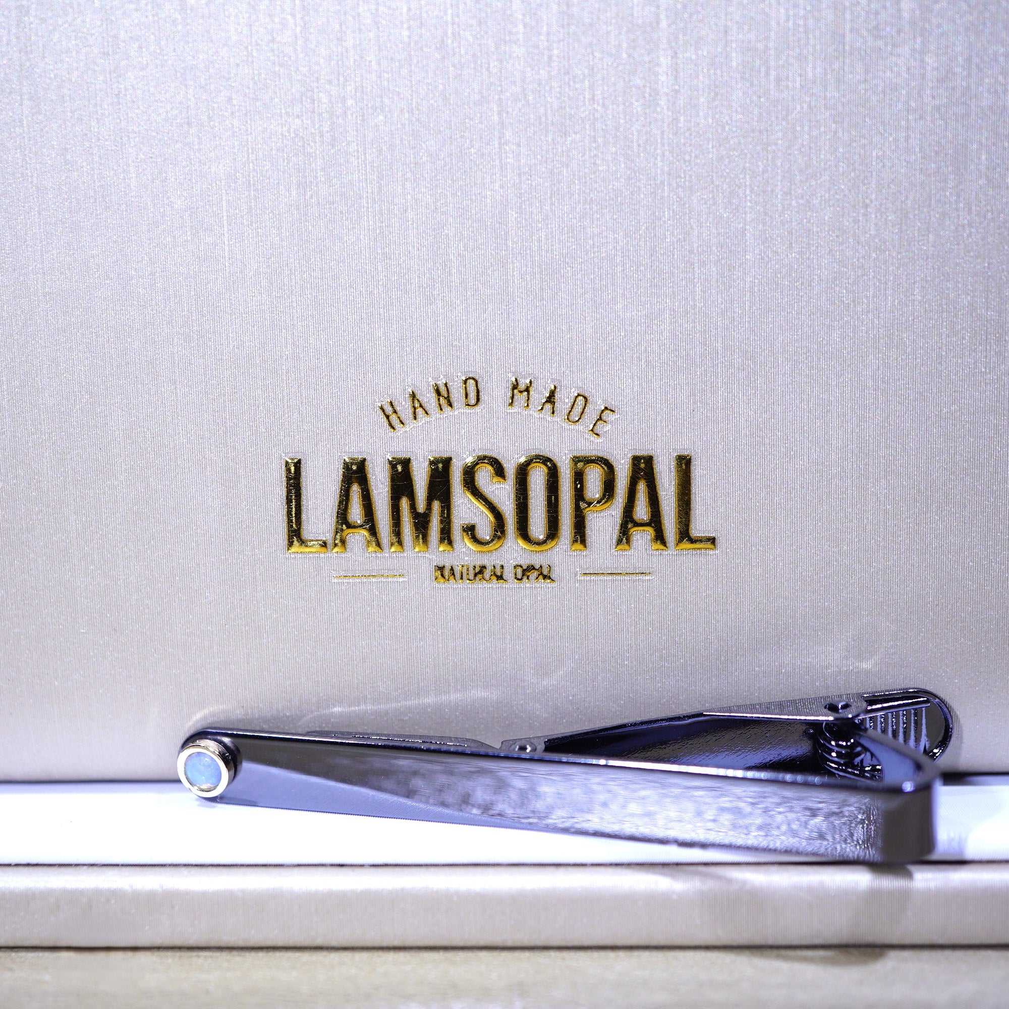 A silver metal tie clip with a textured finish, resting on a white surface in front of a textured light gray background with gold foil branding that reads 'LAMSOPAL HAND MADE'.