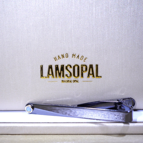 A silver metal tie clip with a textured finish, resting on a white surface in front of a textured light gray background with gold foil branding that reads 'LAMSOPAL HAND MADE'.