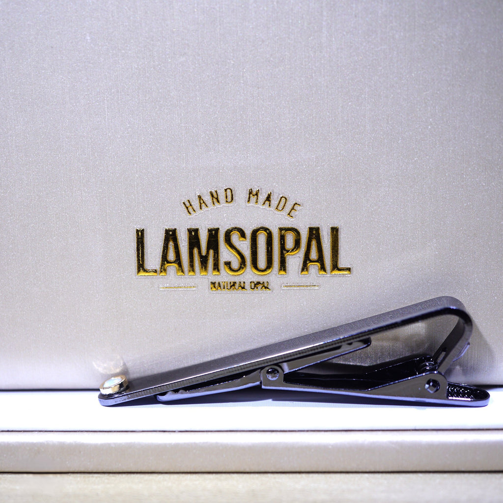 A black metal tie clip resting on a light beige textured surface in front of a matching beige background card printed with the gold and black 'LAMSOPAL HAND MADE NATURAL DOPIA' logo.