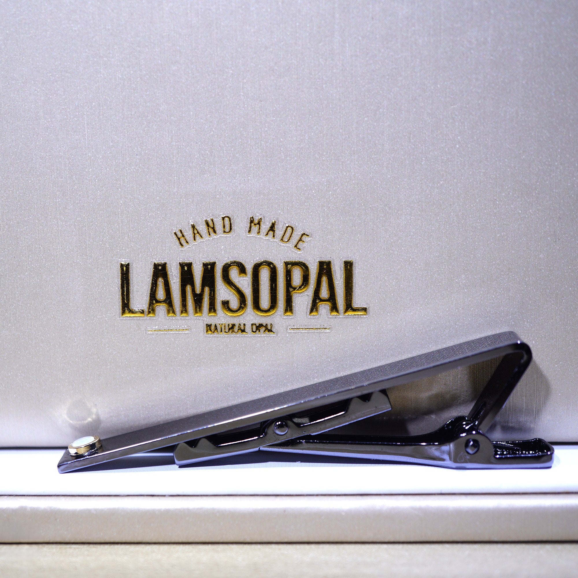 A black metal hair clipper resting on a white surface in front of a textured light gray background with the brand name 'LAMSOPAL' printed in gold.