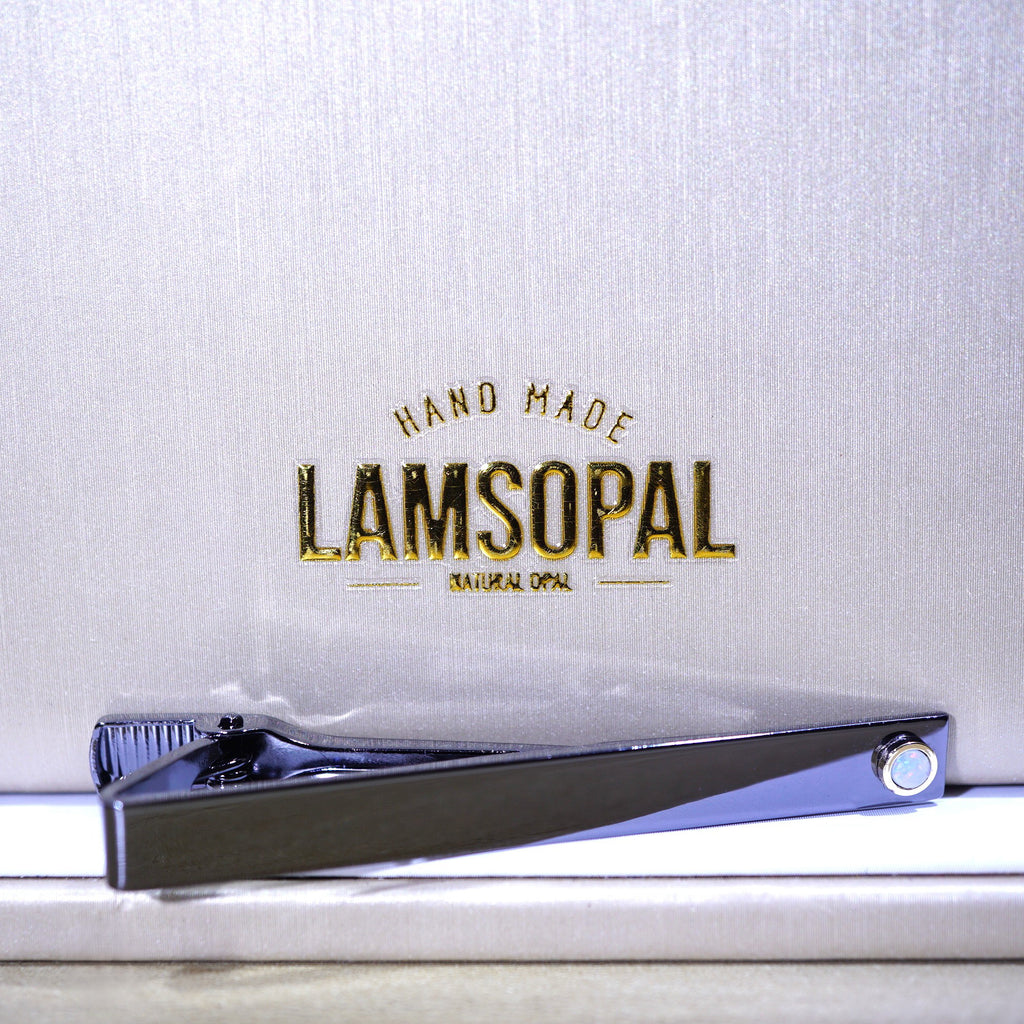 A polished silver tie clip resting on a white surface in front of a textured silver backdrop with gold foil branding that reads 'HAND MADE LAMSOPAL NATURAL OPAL'.