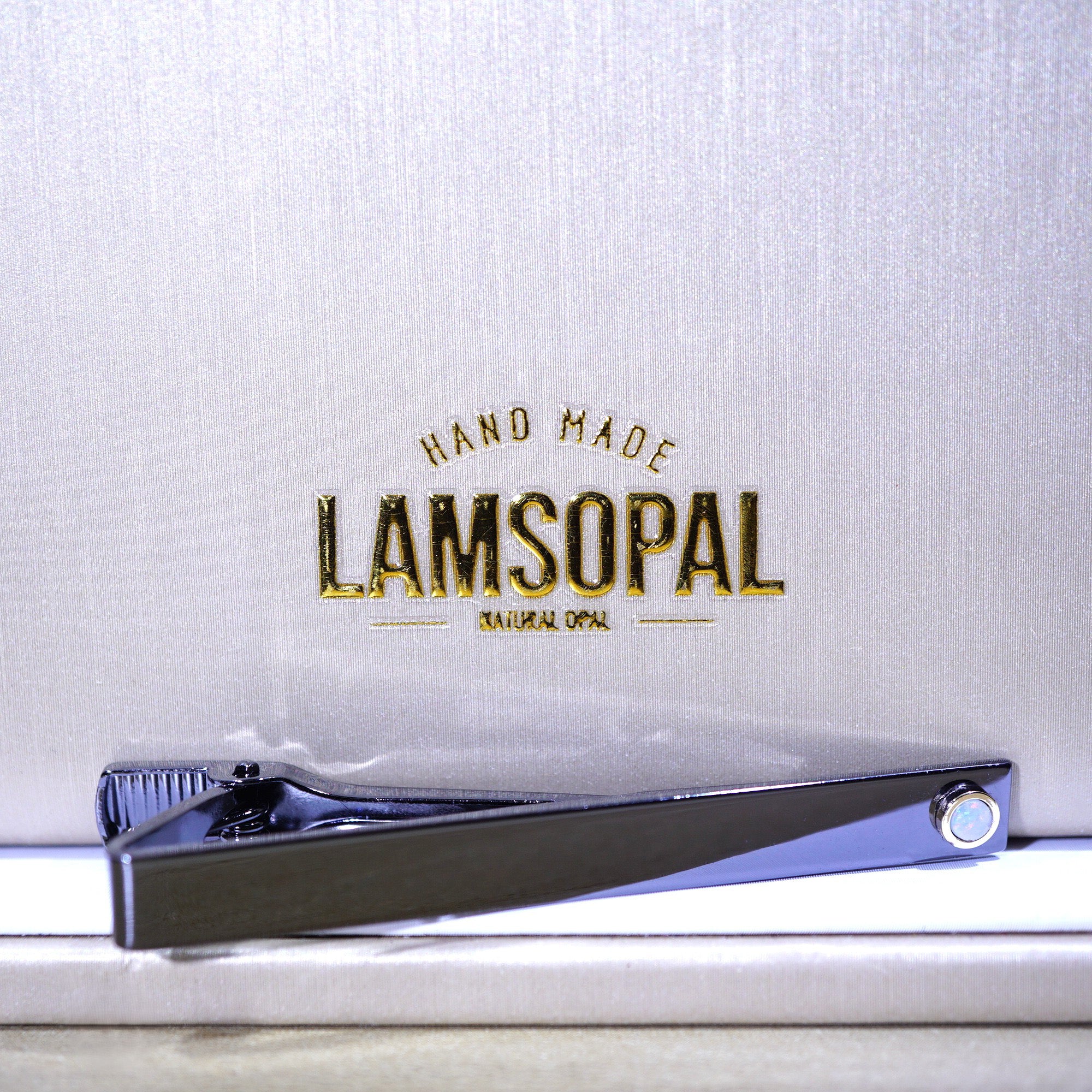 A polished silver tie clip resting on a white surface in front of a textured silver backdrop with gold foil branding that reads 'HAND MADE LAMSOPAL NATURAL OPAL'.