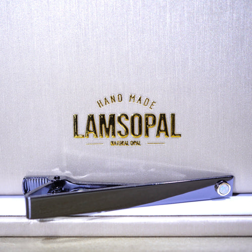 A polished silver tie clip resting on a white surface in front of a textured silver backdrop with gold foil branding that reads 'HAND MADE LAMSOPAL NATURAL OPAL'.