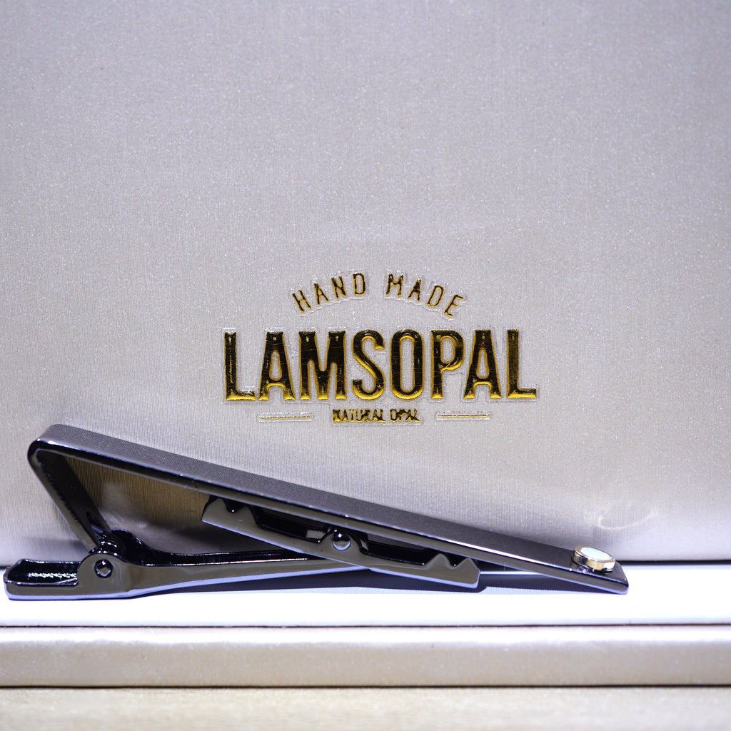 A black metal tie clip with a small silver accent, resting on a light beige textured surface in front of a matching beige background card featuring the gold embossed brand name 'LAMSOPAL'.