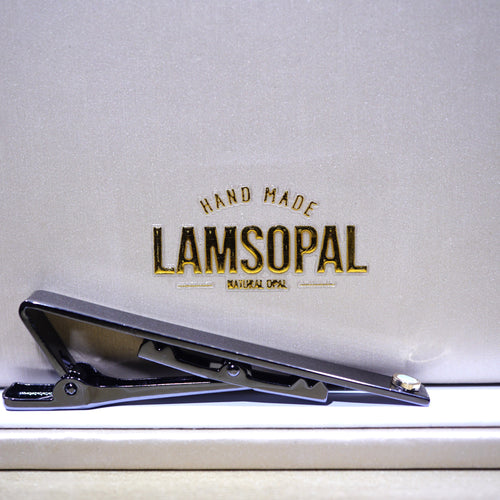 A black metal tie clip with a small silver accent, resting on a light beige textured surface in front of a matching beige background card featuring the gold embossed brand name 'LAMSOPAL'.