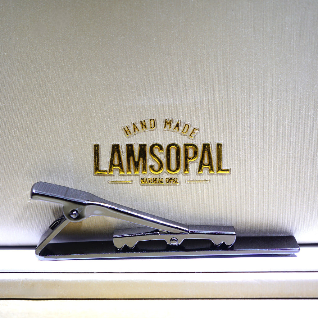 A silver-toned metal tie clip resting on a beige textured surface with gold embossed branding that reads 'HAND MADE LAMSOPAL'.