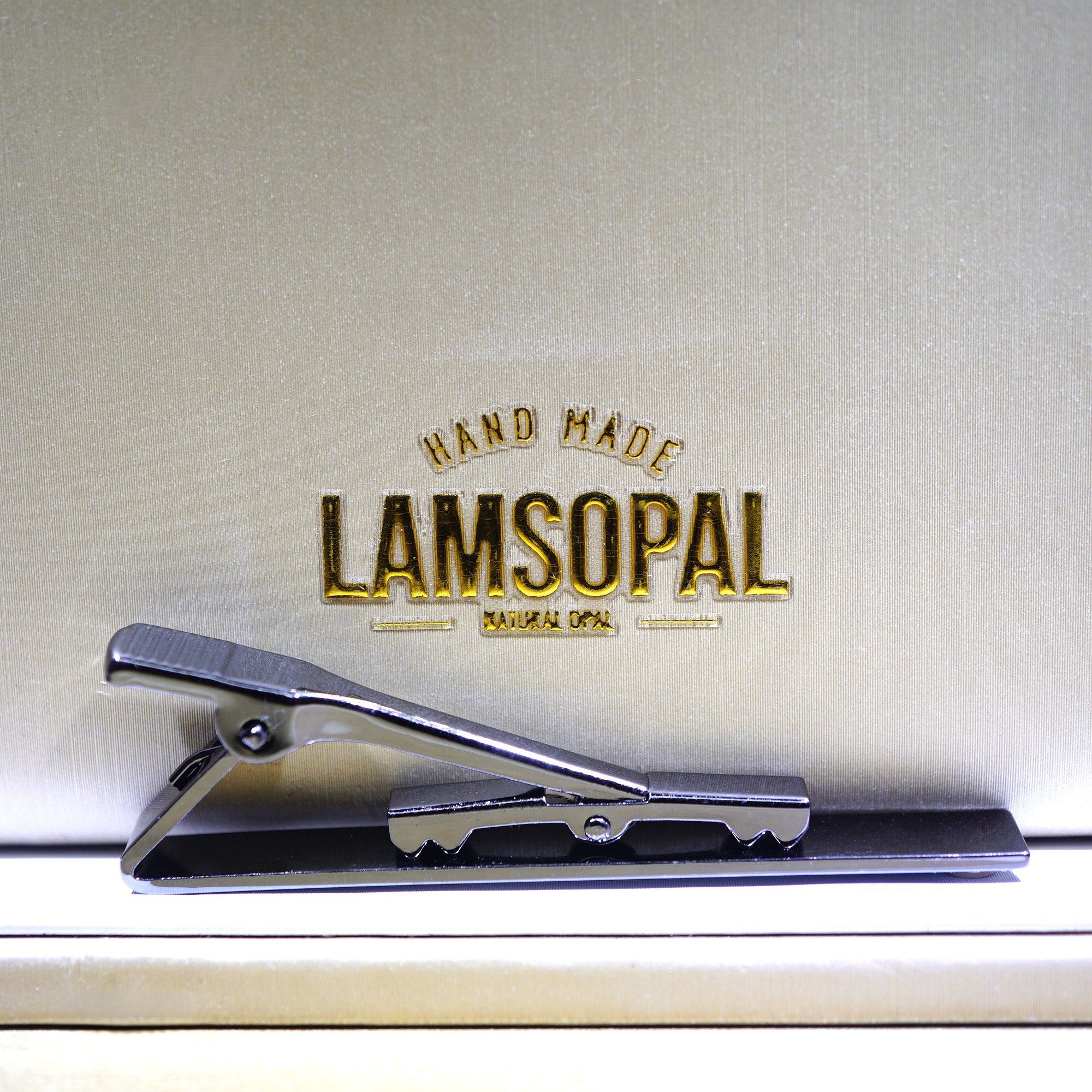 A silver-toned metal tie clip resting on a beige textured surface with gold embossed branding that reads 'HAND MADE LAMSOPAL'.