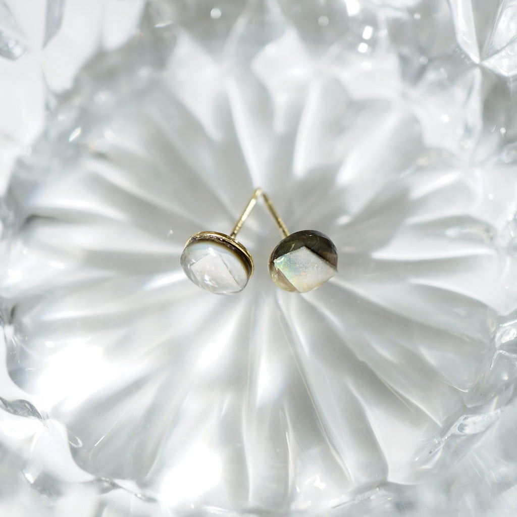 A pair of gold stud earrings with oval cabochon gemstones, one clear and one iridescent white, resting on a textured clear glass surface.