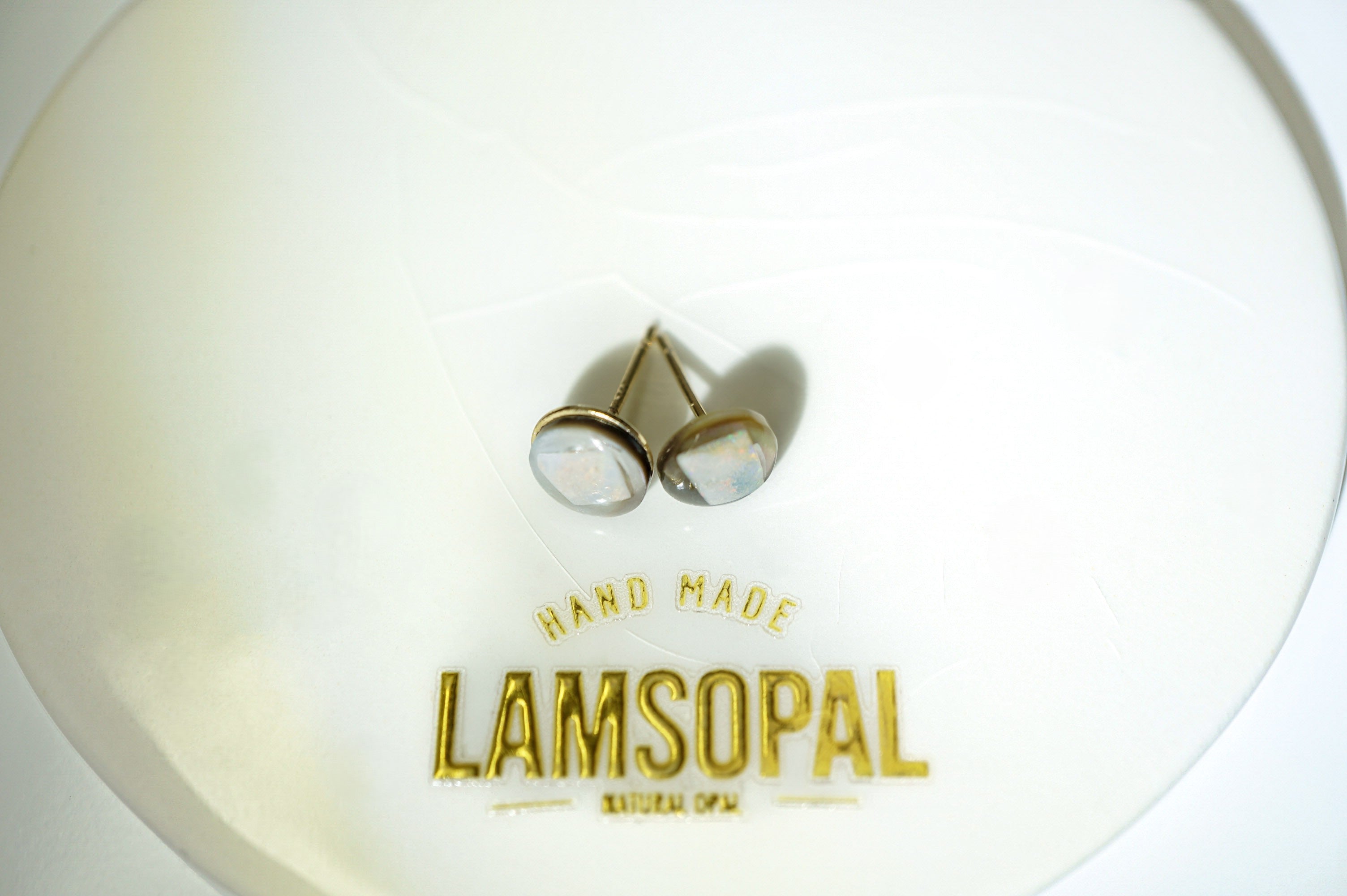 A pair of gold-toned stud earrings with round, iridescent shell-like cabochons resting on a white circular display card printed with the brand name 'LAMSOPAL'.