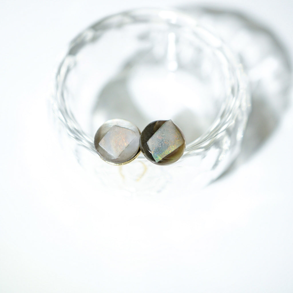 Two faceted oval gemstones with a metallic gold setting, displayed inside a clear textured glass bowl on a bright white background.