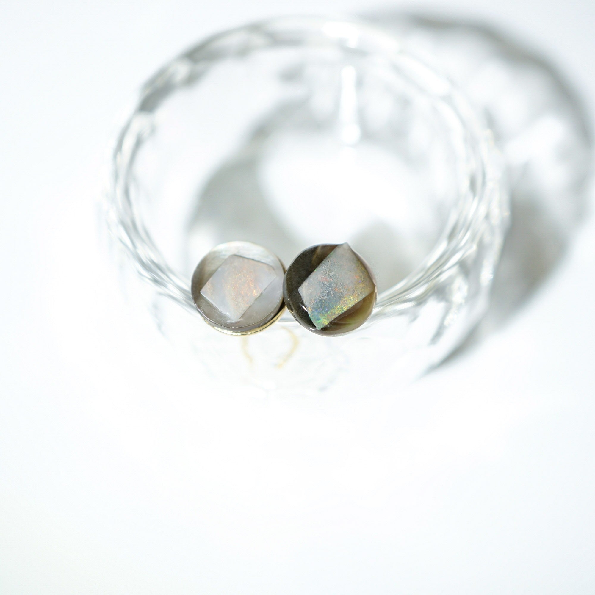 Two faceted oval gemstones with a metallic gold setting, displayed inside a clear textured glass bowl on a bright white background.