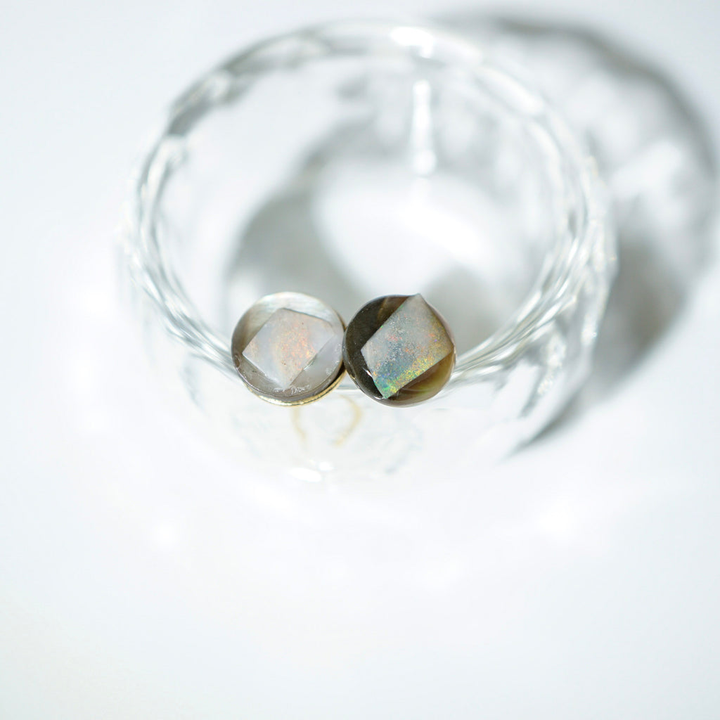 Two faceted oval gemstone beads with a smoky gray-brown body and iridescent rainbow flashes, displayed inside a clear glass bowl on a white background.