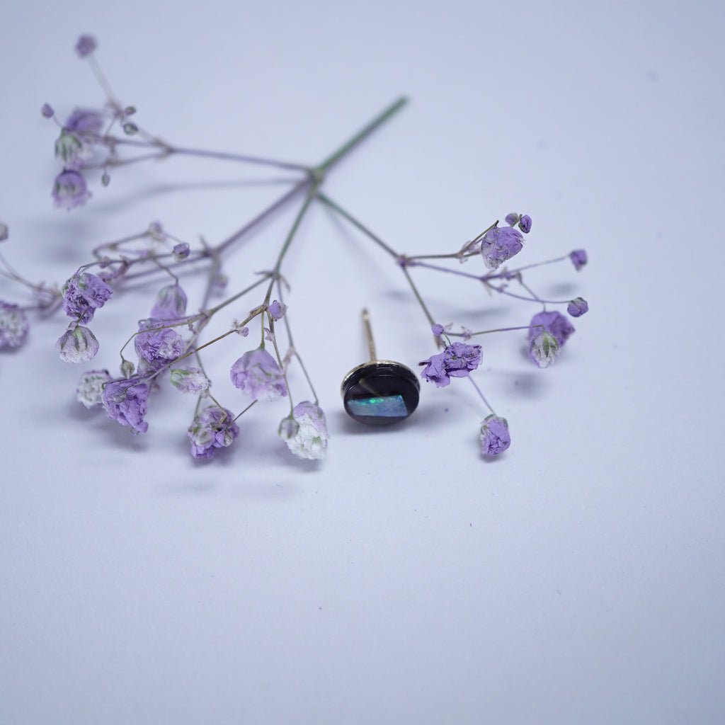 A single black oval stud earring with an iridescent blue-green stone, displayed on a light lavender background with dried purple flowers as decorative props.