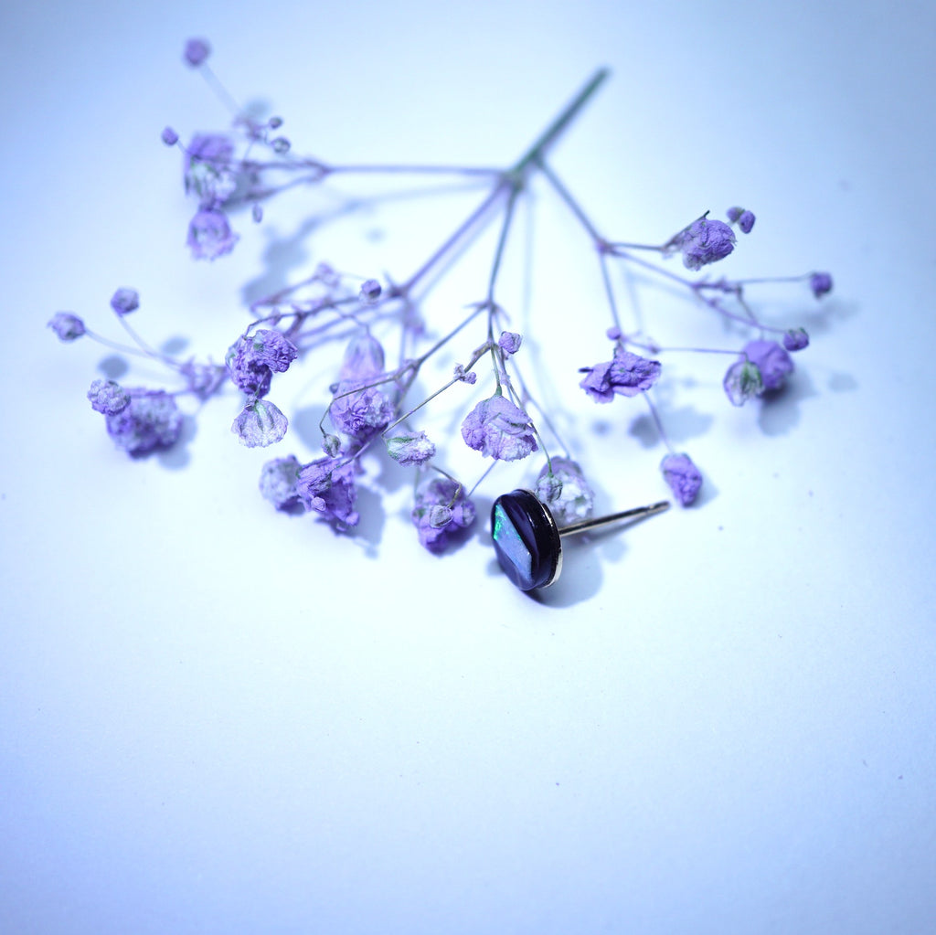 A single earring with a faceted dark purple oval gemstone in a silver-toned setting, resting on delicate dried lavender sprigs against a pale blue-white background.