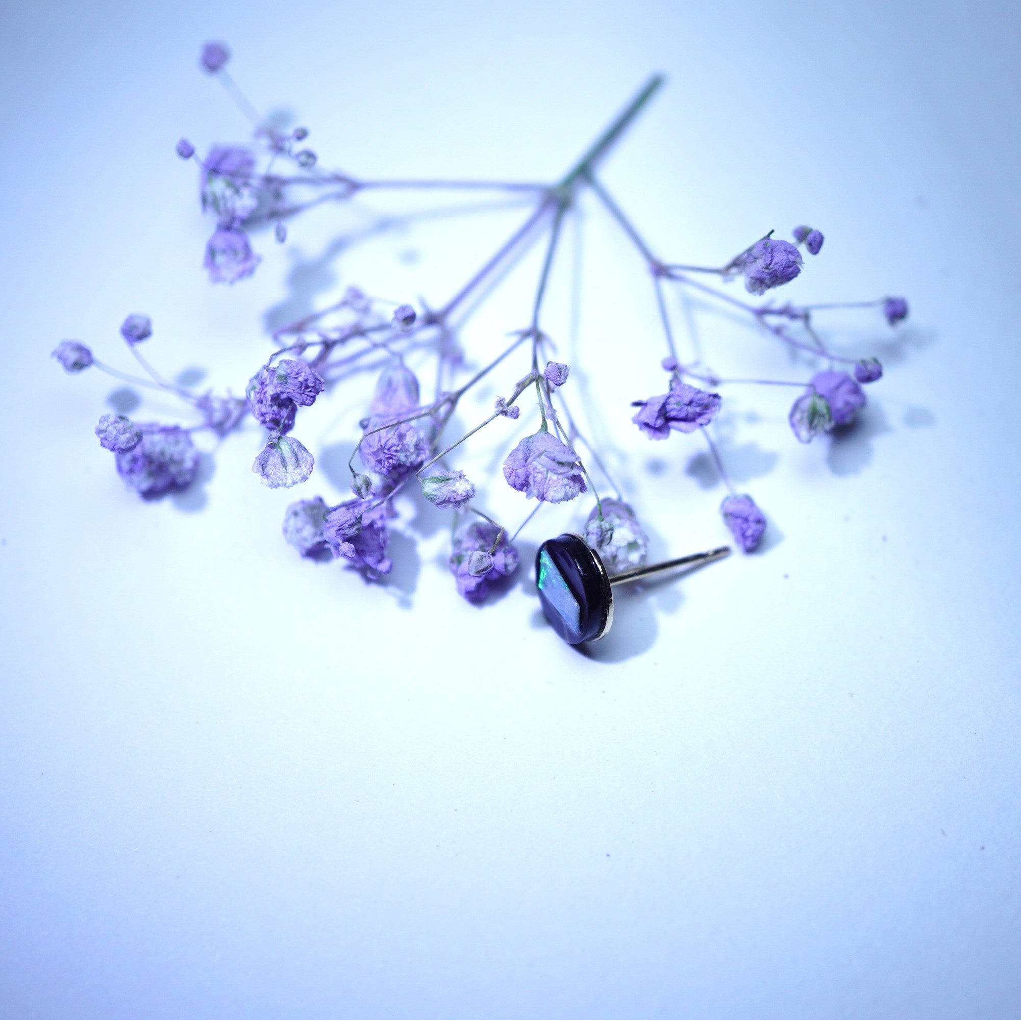 A single earring with a faceted dark purple oval gemstone in a silver-toned setting, resting on delicate dried lavender sprigs against a pale blue-white background.