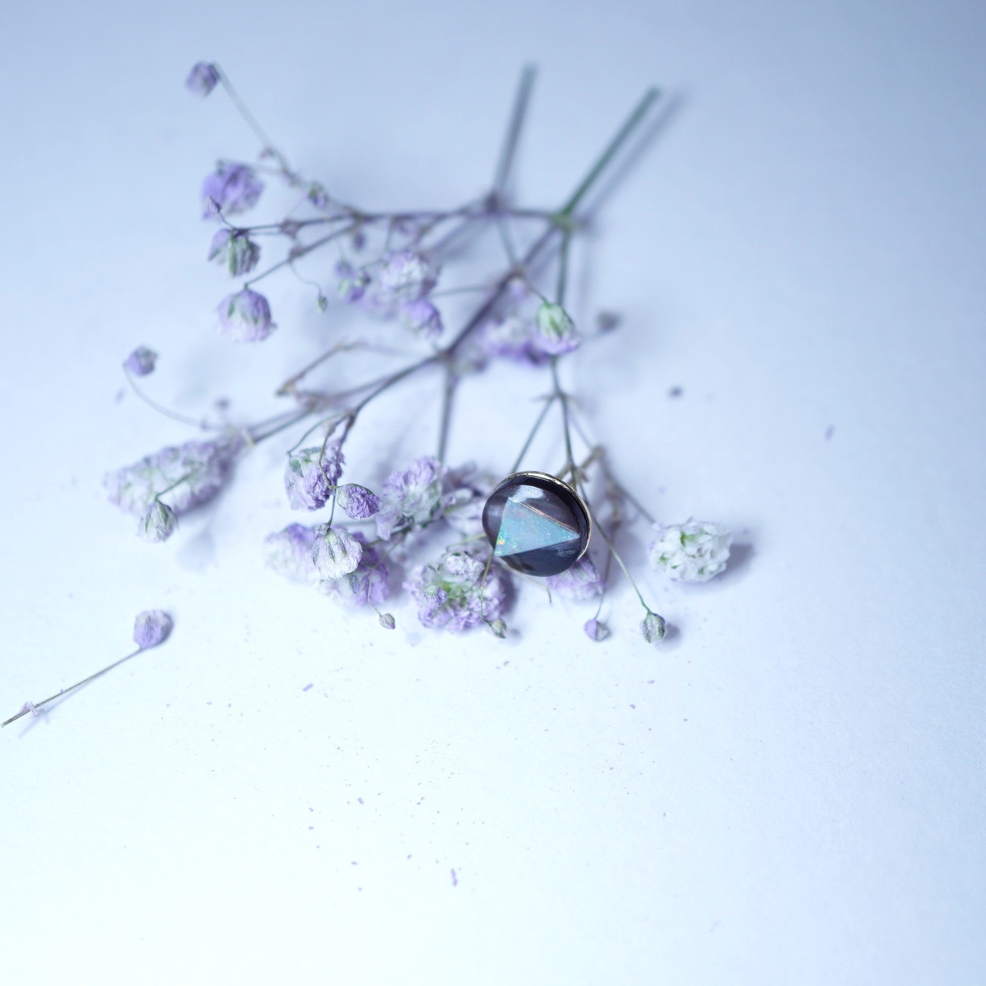 A single round bezel-set gemstone earring with a dark iridescent blue-green center and silver-toned setting, styled with dried lavender flowers on a pale lavender background.