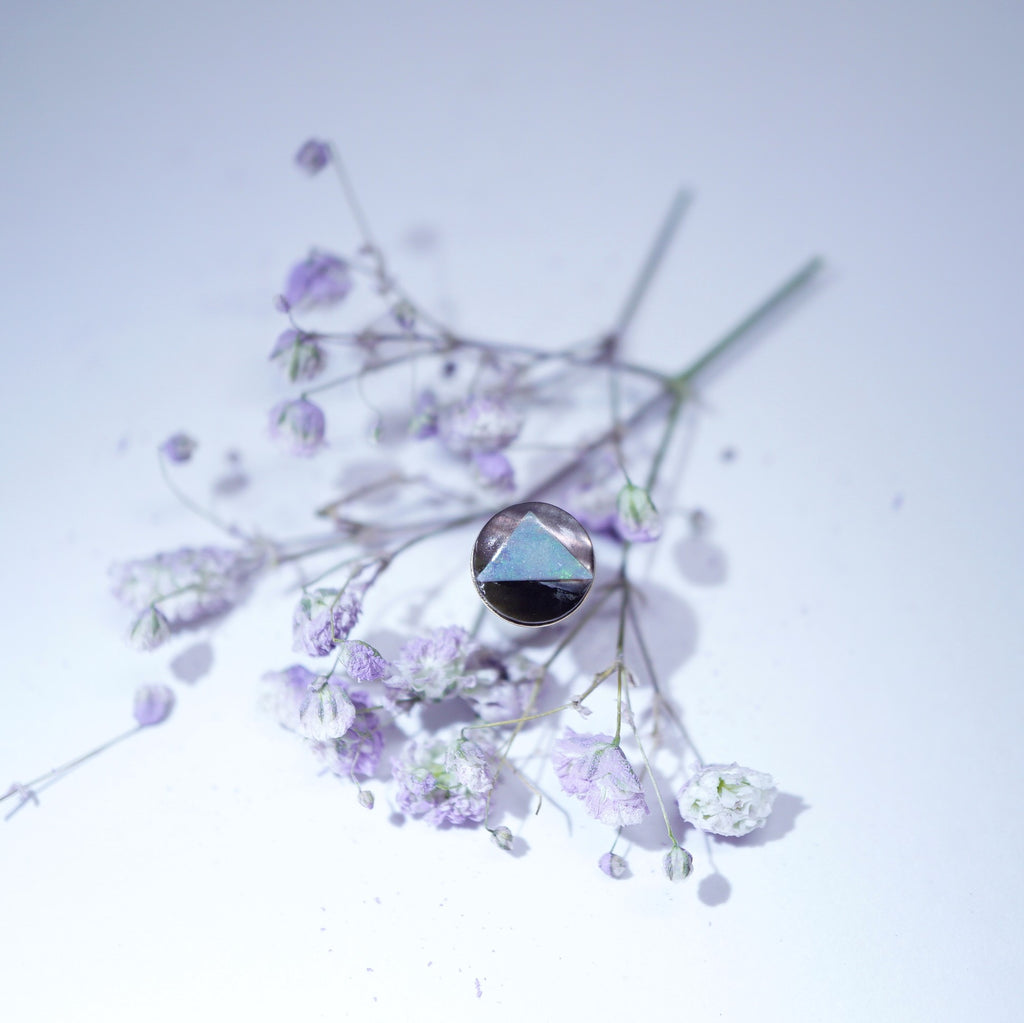 A single round stud earring with a faceted iridescent blue-green gemstone in a dark bezel setting, artistically placed among dried lavender and white baby's breath flowers on a light, cool-toned background.